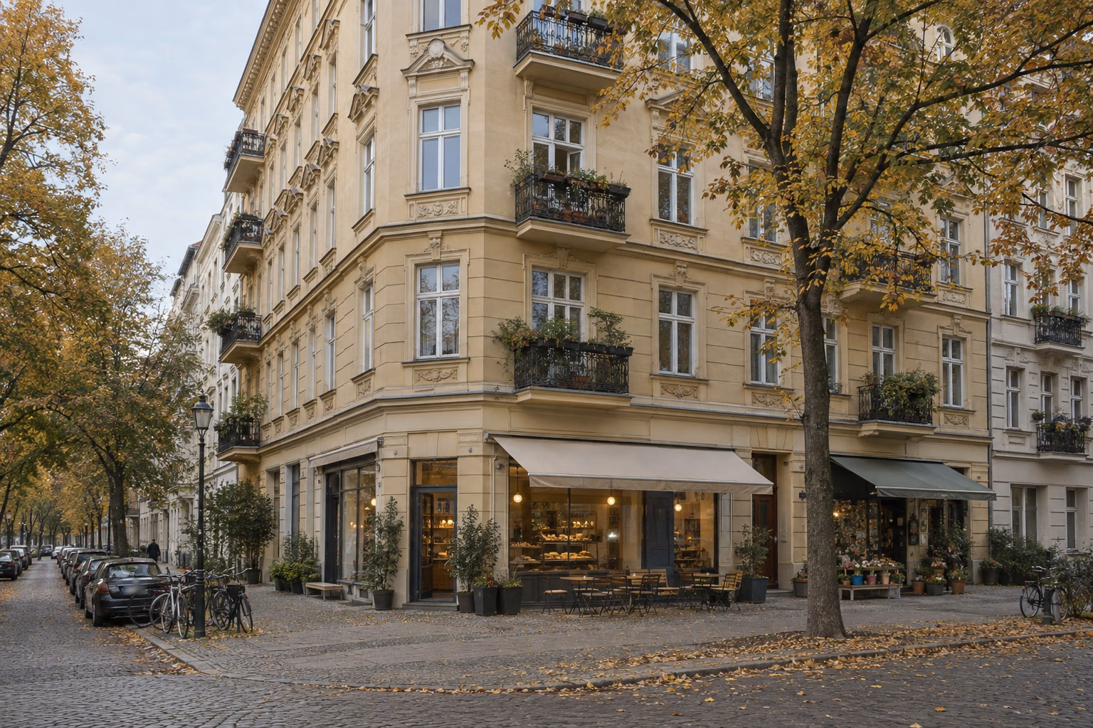 Berliner Altbau-Eckhaus mit Bäckerei und Laden im Erdgeschoss und Wohnungen mit Balkonen darüber, an einer ruhigen Straße mit Herbstbäumen.