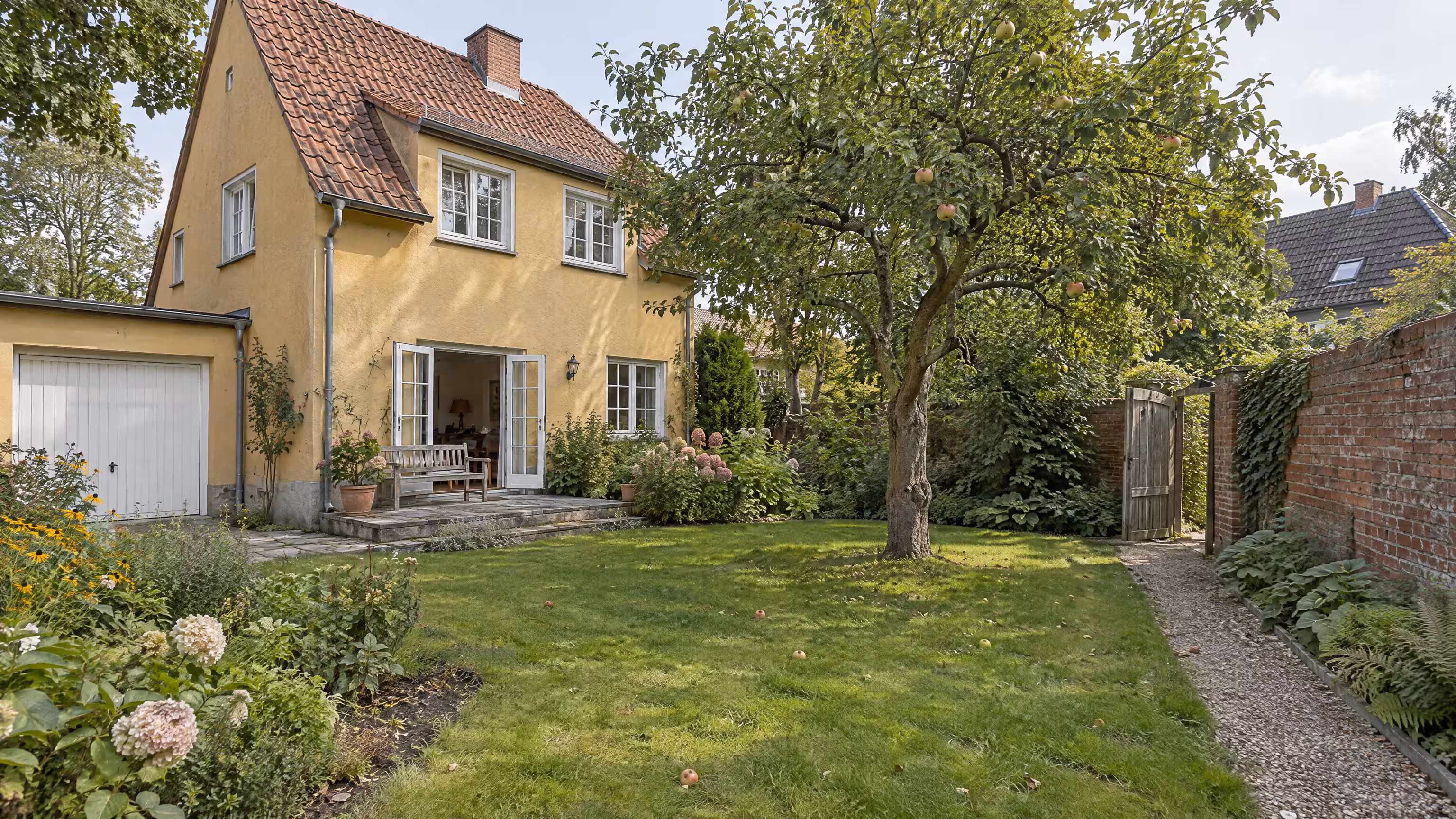 Illustrative Gartenseite eines älteren Wohnhauses mit Terrasse und Garage, Apfelbaum im Garten und ein Kiesweg zum Gartentor in der Backsteinmauer.