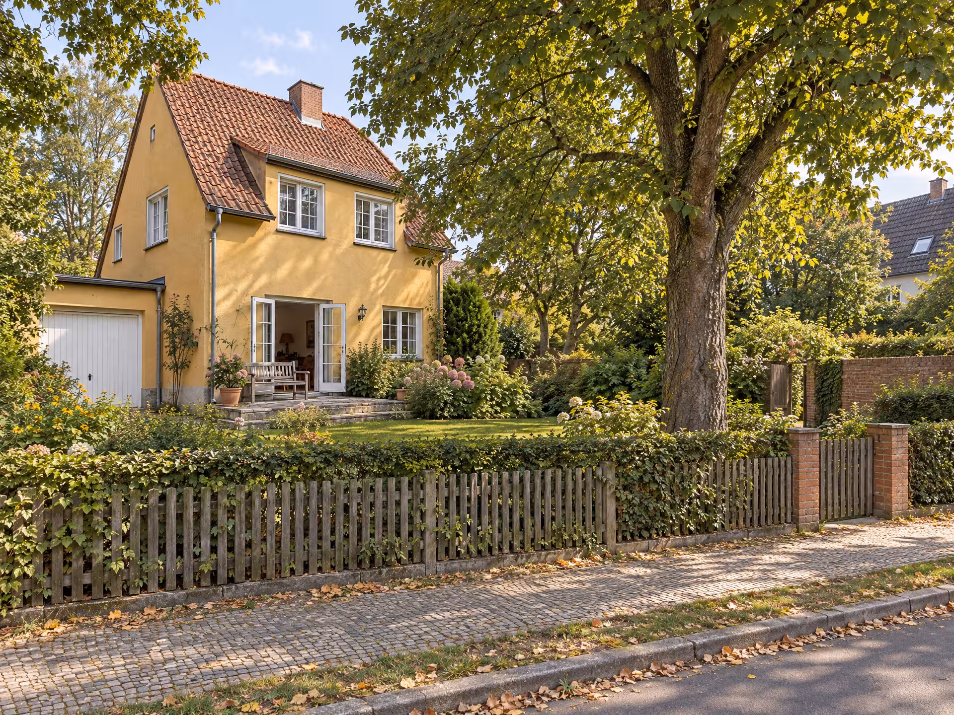 Illustratives Motiv: ein Wohnhaus mit Vorgarten hinter Holzzaun und Hecke an einer ruhigen Straße.