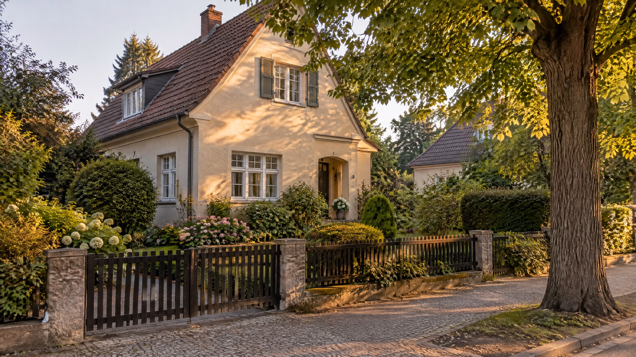 Illustratives Motiv: Ein freistehendes Einfamilienhaus aus den 1930er-Jahren mit Vorgarten, Holzzaun und einer Linde im warmen Abendlicht.
