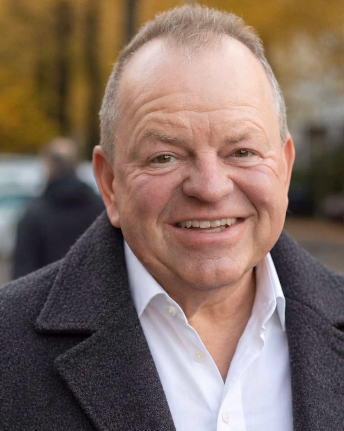 Oliver Preikschat lächelt, im grauen Mantel auf einer Berliner Straße im Herbst.