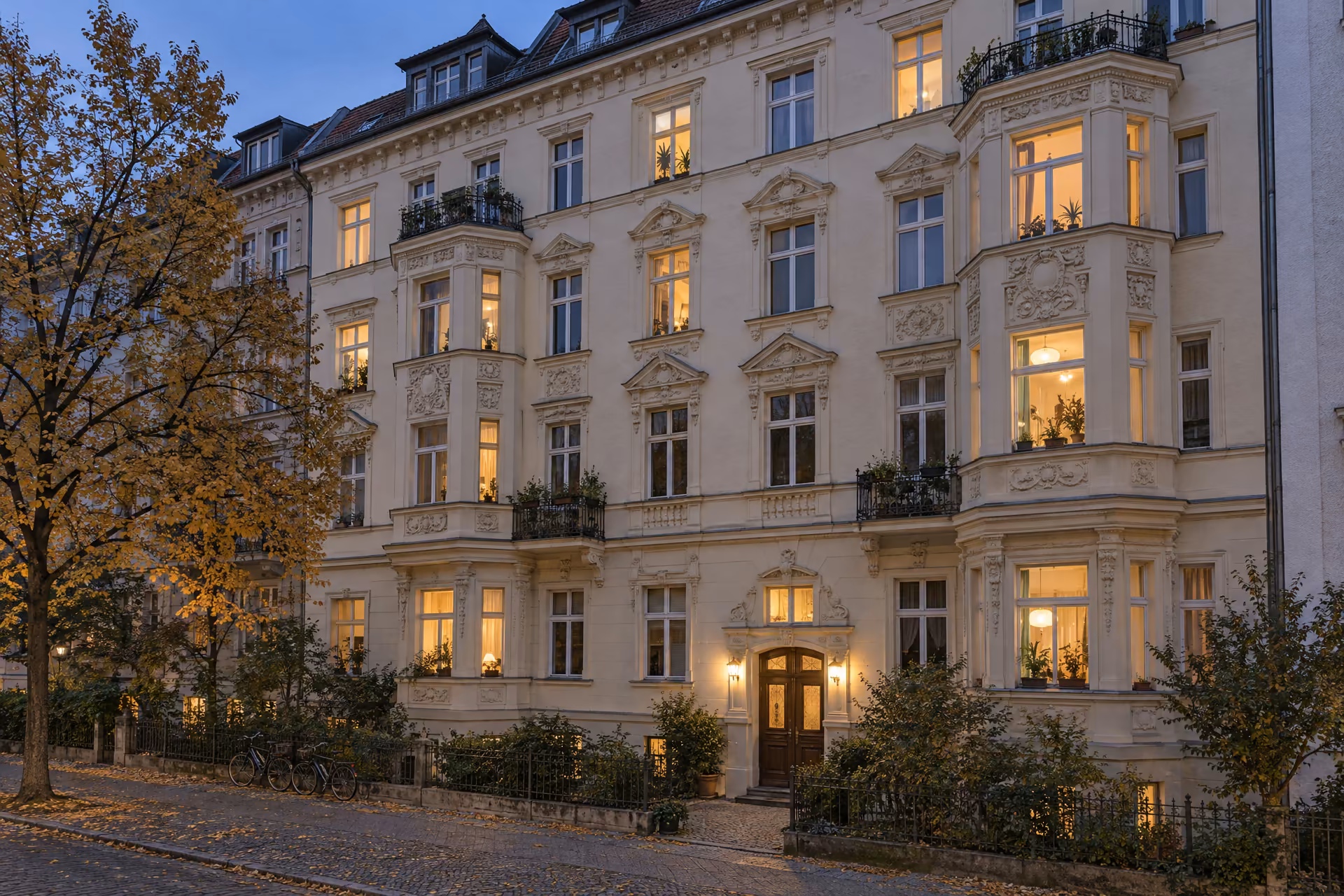 Gründerzeit-Mietshaus in Berlin-Friedenau zur blauen Stunde: helle Stuckfassade mit zwei Erkern und kleinen Balkonen, hinter vielen Fenstern brennt unterschiedlich warmes Licht, davor ein Herbstbaum und Fahrräder am Zaun.