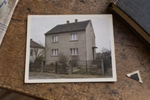 Leicht verblasster Farbabzug aus den 1990er-Jahren auf einem Holztisch. Er zeigt ein zweigeschossiges Haus mit grauem Putz hinter einem Drahtzaun.