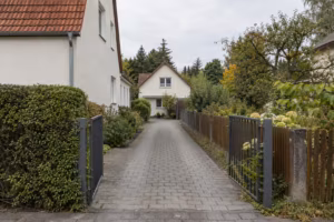 Gepflasterte Zufahrt zwischen einem verputzten Einfamilienhaus mit Hecke und einem Holzzaun. Am Ende der Zufahrt steht ein zweites, kleineres Haus auf dem hinteren Grundstück.