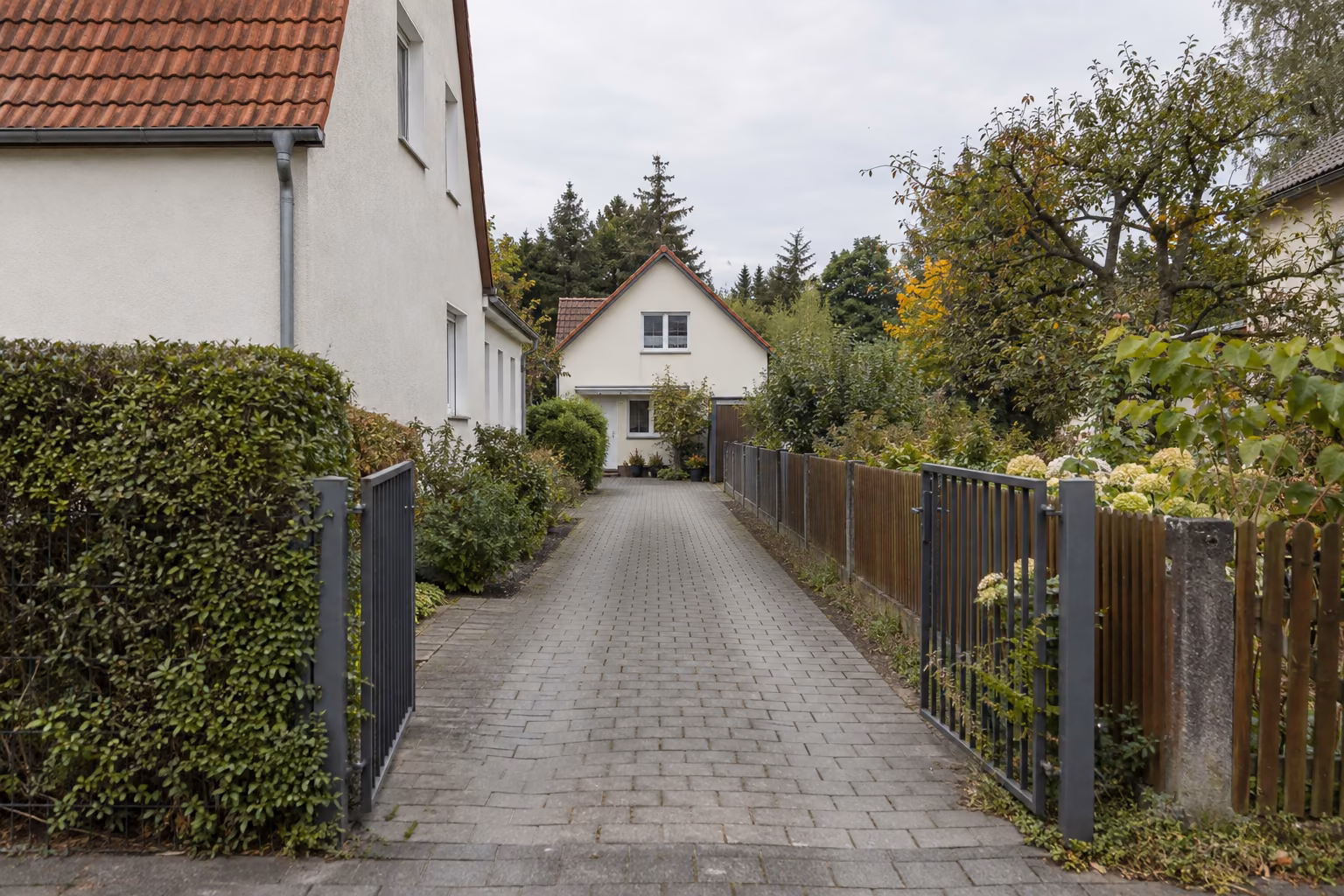 Gepflasterte Zufahrt zwischen einem verputzten Einfamilienhaus mit Hecke und einem Holzzaun. Am Ende der Zufahrt steht ein zweites, kleineres Haus auf dem hinteren Grundstück.