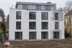 Rückseite eines neuen, viergeschossigen Mehrfamilienhauses mit weißem Putz und französischen Balkonen; ein Teil der Fassade ist nur grau verputzt, davor liegt ein unbepflanzter Garten mit gestapeltem Holz.