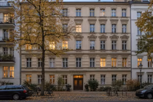 Fassade eines fünfgeschossigen Berliner Mietshauses der Gründerzeit im Abendlicht. Einige Fenster sind erleuchtet, davor steht ein Straßenbaum mit gelbem Laub.