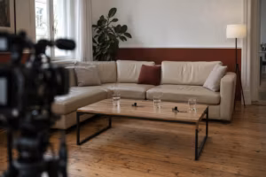 Helles Ecksofa in einem Altbaubüro vor dem Interview. Auf dem Tisch davor stehen drei Gläser Wasser und liegen zwei Ansteckmikrofone, vorn unscharf eine Kamera auf einem Stativ.