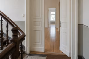 Halb geöffnete weiße Flügeltür einer Altbauwohnung, im Schloss steckt ein Schlüssel. Dahinter liegt ein heller, leerer Flur mit Holzdielen.