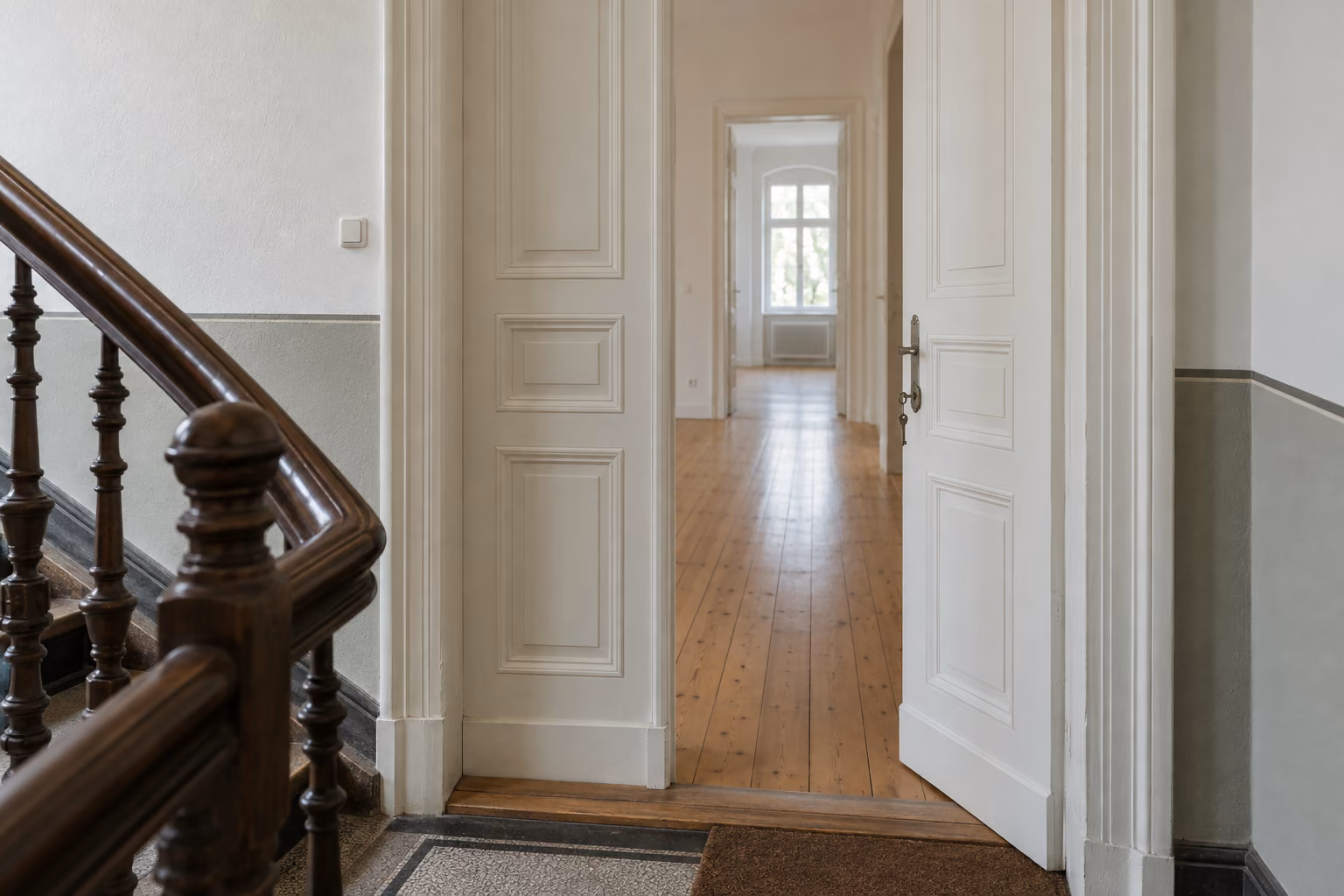 Halb geöffnete weiße Flügeltür einer Altbauwohnung, im Schloss steckt ein Schlüssel. Dahinter liegt ein heller, leerer Flur mit Holzdielen.