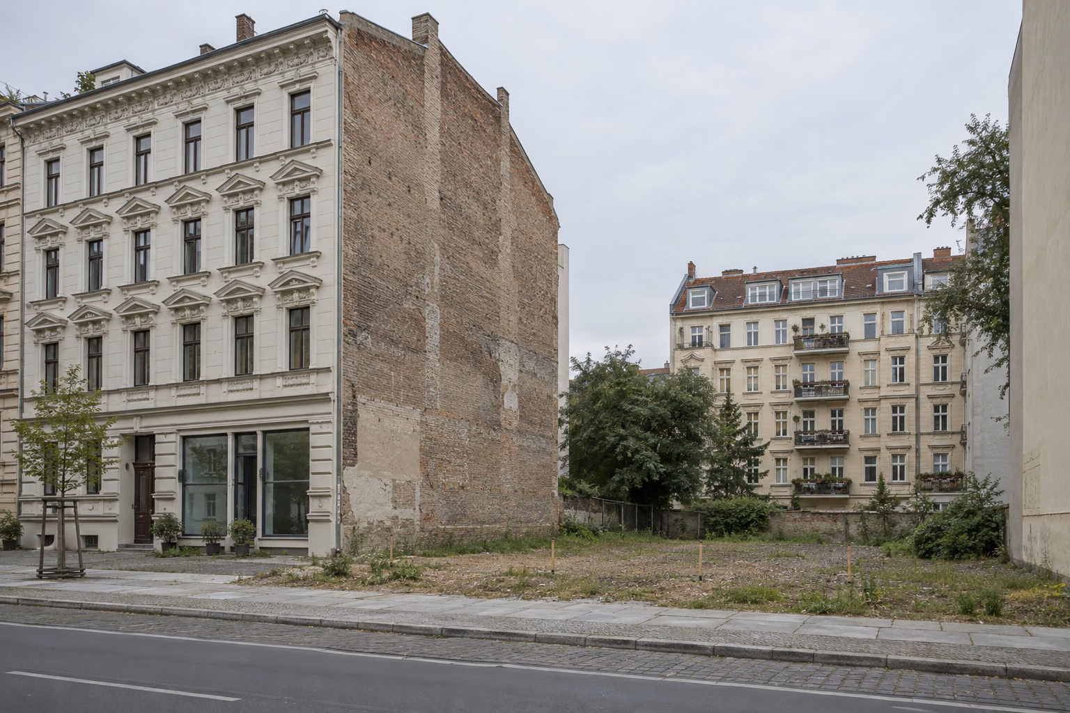 Fünfgeschossiges Gründerzeithaus mit heller Stuckfassade, dessen unverputzte Ziegel-Brandwand an ein leeres, mit Pflöcken abgestecktes Grundstück grenzt; dahinter Bäume und ein weiteres Altbau-Wohnhaus unter grauem Himmel.