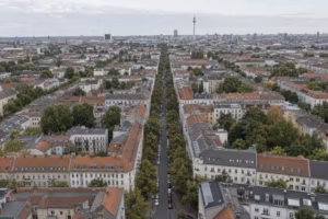 Blick von oben auf ein Berliner Wohnquartier mit Blockrandbebauung, roten Dächern und einer baumbestandenen Straße. Am Horizont steht der Fernsehturm.