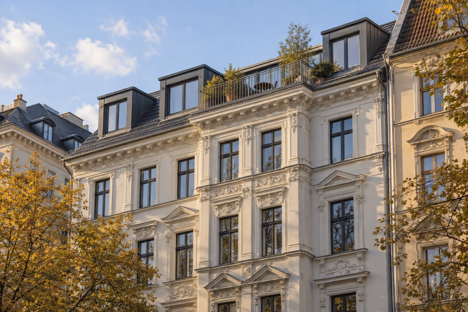 Gründerzeithaus mit heller Stuckfassade, Dachgauben und kleiner Dachterrasse im Abendlicht, davor gelbes Herbstlaub.