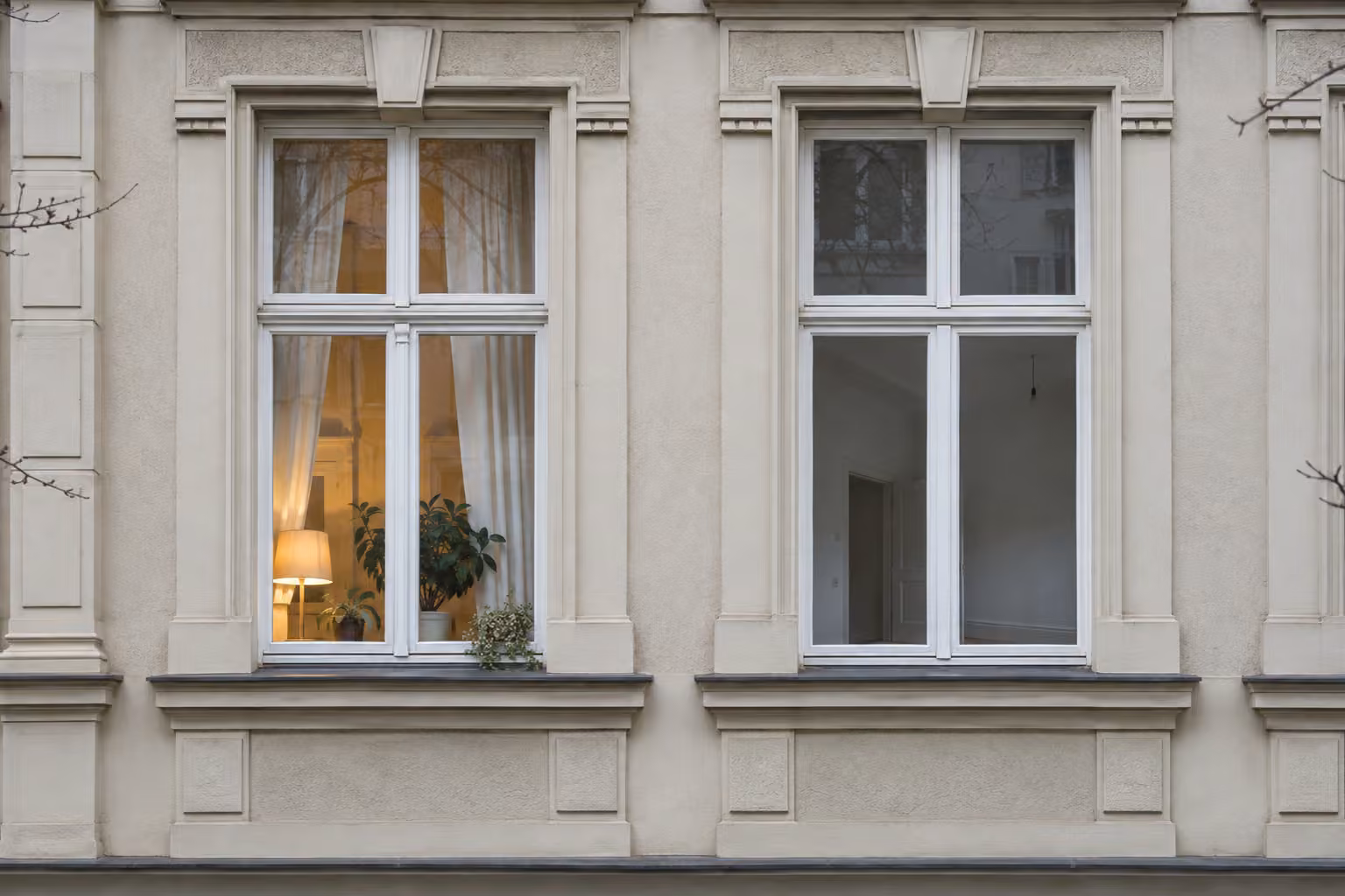 Helle Altbaufassade mit zwei großen Fenstern; hinter einem Fenster ist ein eingerichteter Wohnraum zu sehen.