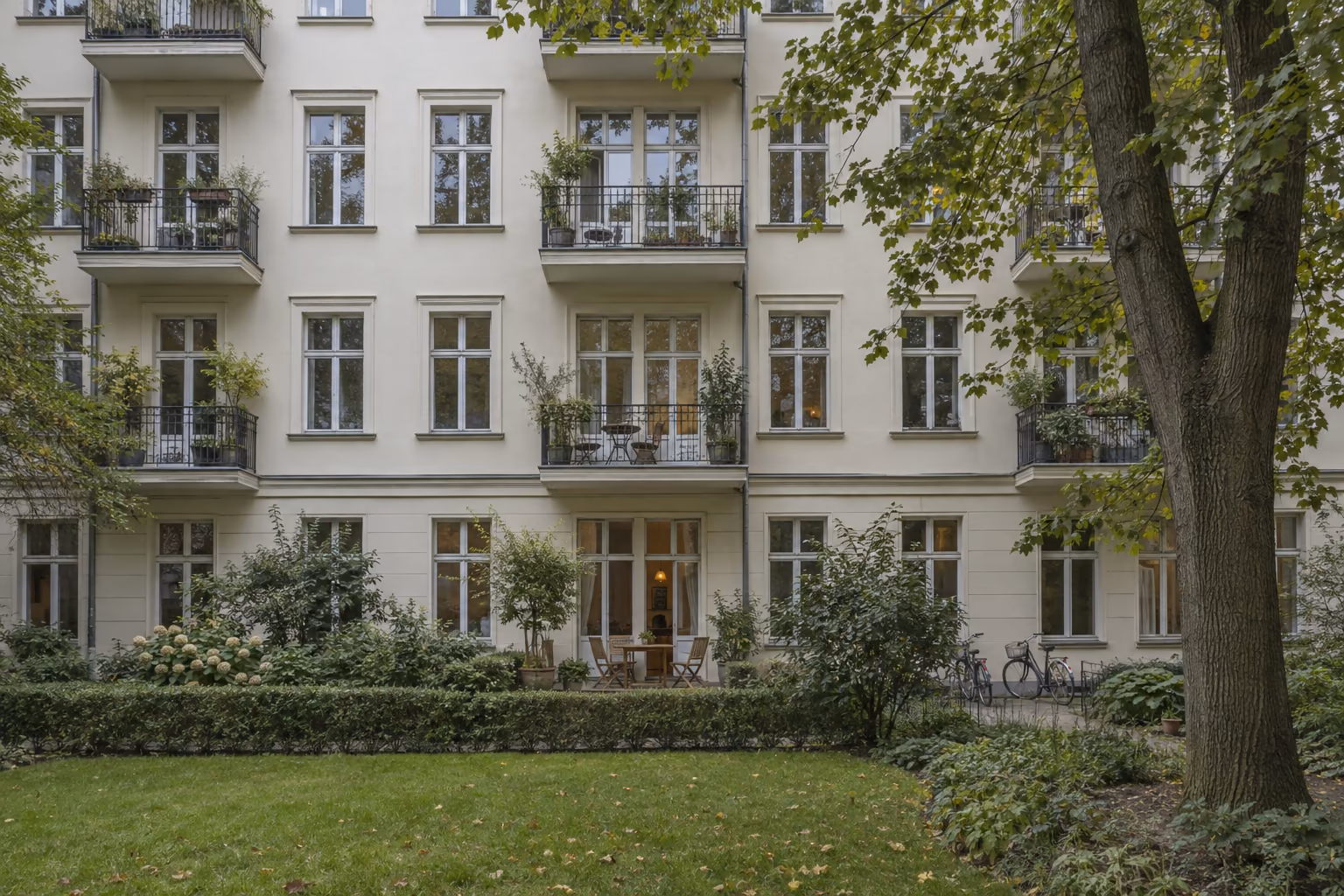 Gartenseite eines hell verputzten Berliner Mietshauses mit Balkonen. Im Erdgeschoss hat eine Wohnung eine eigene kleine Terrasse mit Tisch und Stühlen, die eine niedrige Hecke vom gemeinsamen Rasen trennt.