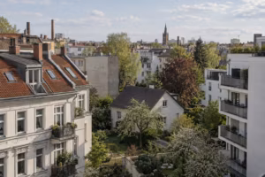 Blick aus einem oberen Stockwerk über ein Berliner Wohnviertel. Vorn stehen Altbauten mit Ziegeldächern, daneben ein kleines Einfamilienhaus mit Garten und ein neuer Wohnblock, dahinter weitere Dächer und ein Kirchturm.