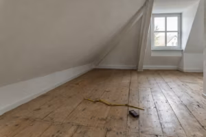 Leeres Dachzimmer mit Dielenboden und steiler Dachschräge. Auf dem Boden liegen ein aufgeklappter Zollstock und ein Laser-Entfernungsmesser.