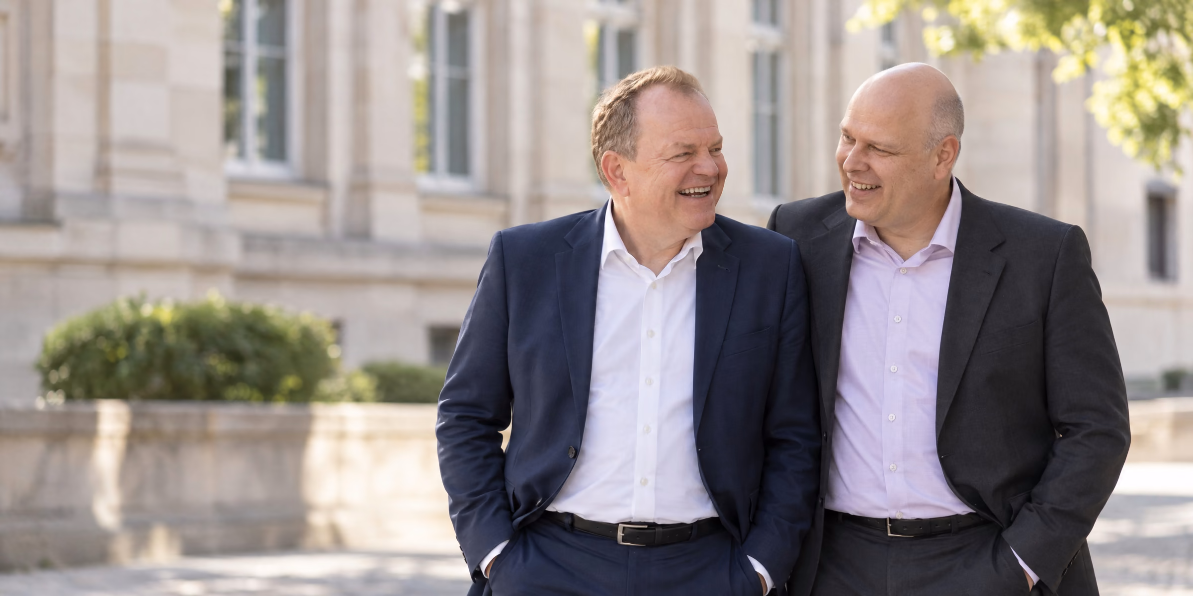 Oliver Preikschat und Mirko Otto stehen nebeneinander vor einem hellen Altbau und lachen miteinander