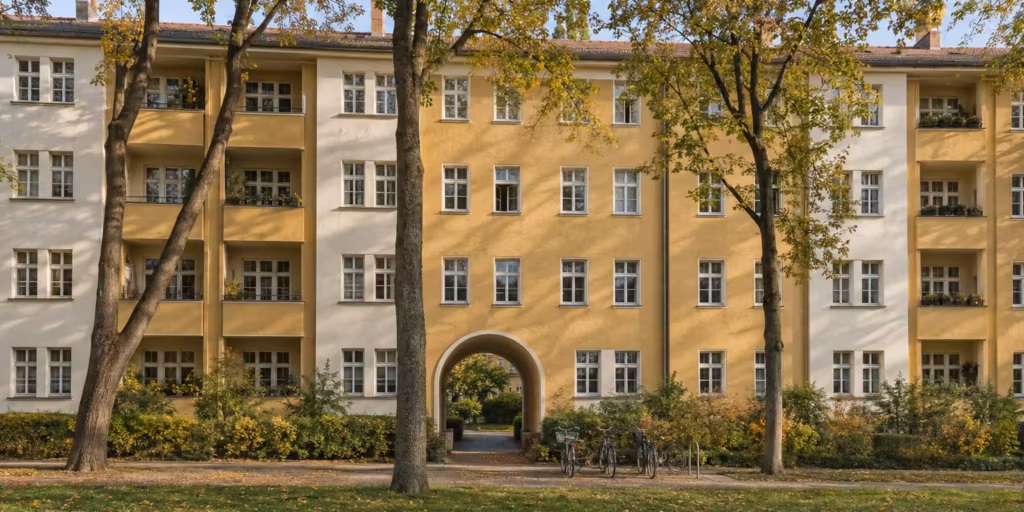 Wohnanlage einer Berliner Genossenschaft aus den 1920er-Jahren: lange Häuserzeile mit ockerfarbenem und weißem Putz, Loggien und einem Torbogen zum begrünten Hof, davor Linden im Herbstlaub.