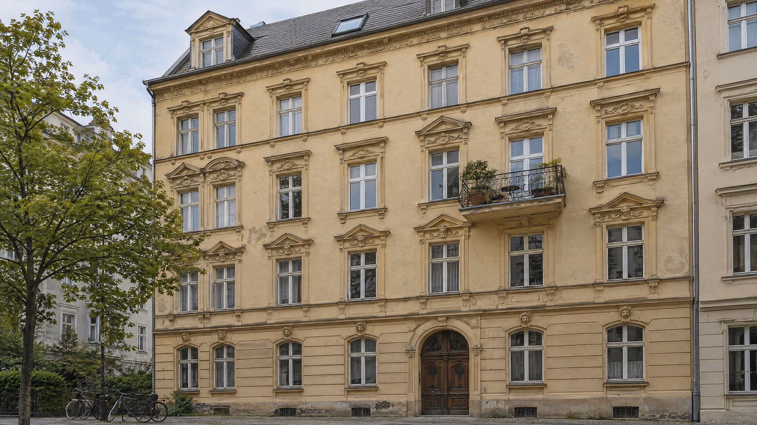 Illustratives Berliner Altbau-Mietshaus mit ockerfarbener Stuckfassade, alten Holzkastenfenstern, einzelnen neueren Fenstern, Balkon und einem neuen Dachfenster im Schieferdach.