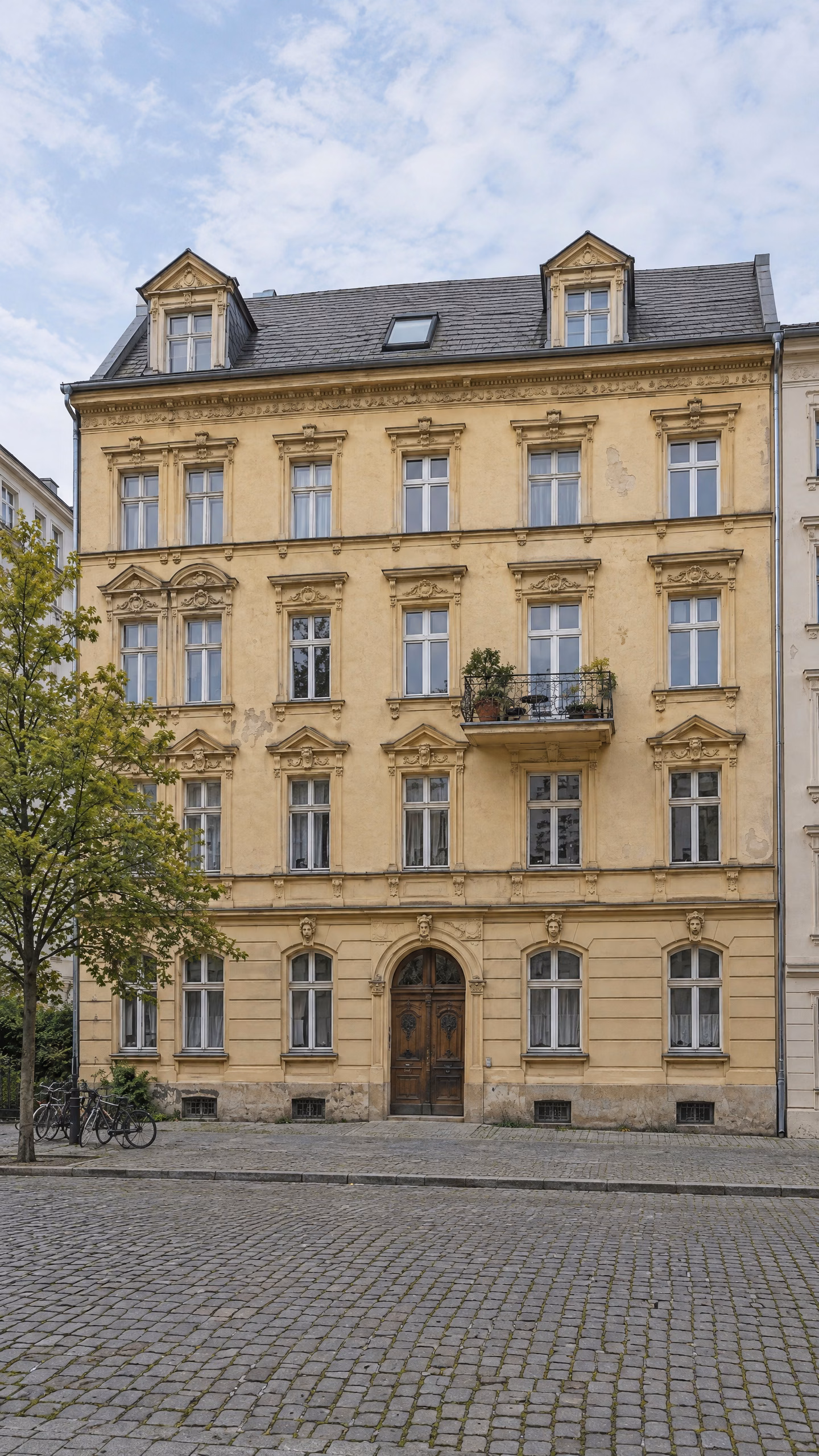 Dasselbe illustrative Mietshaus im Hochformat: Stuckfassade mit alten und neueren Fenstern, Balkon und neues Dachfenster.