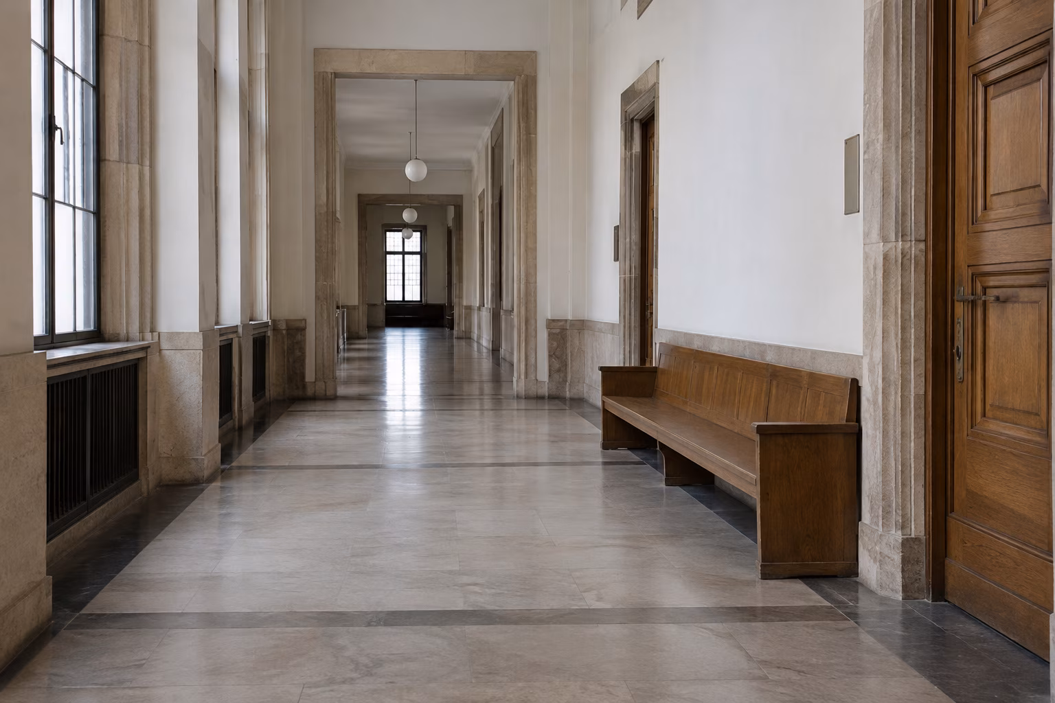 Leerer, heller Gerichtsflur mit Steinboden, hohen Holztüren und einer Holzbank an der Wand.