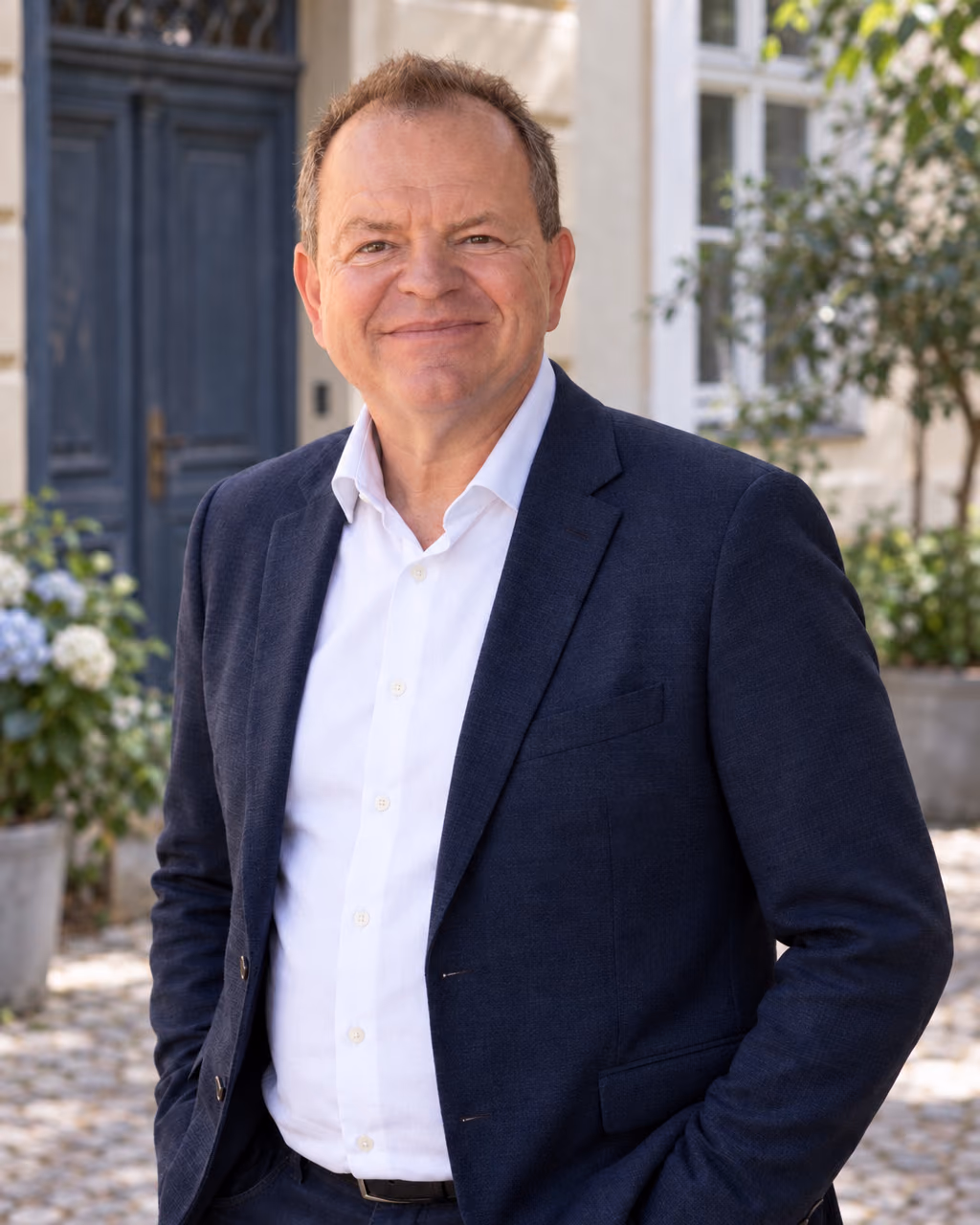 Oliver Preikschat im weißen Hemd und dunkelblauen Sakko vor einer blauen Tür im begrünten Innenhof.