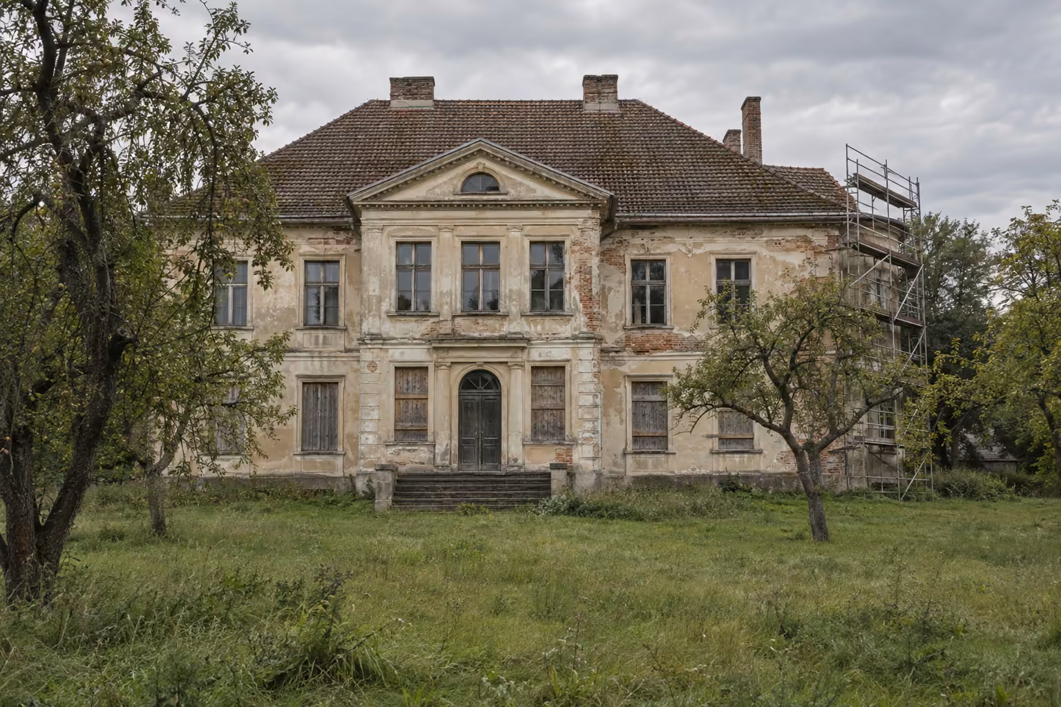 Leerstehendes Gutshaus in Brandenburg mit abblätterndem Putz, mit Brettern verschlossenen Fenstern und einem Gerüst an einer Ecke, davor eine Wiese mit Obstbäumen.