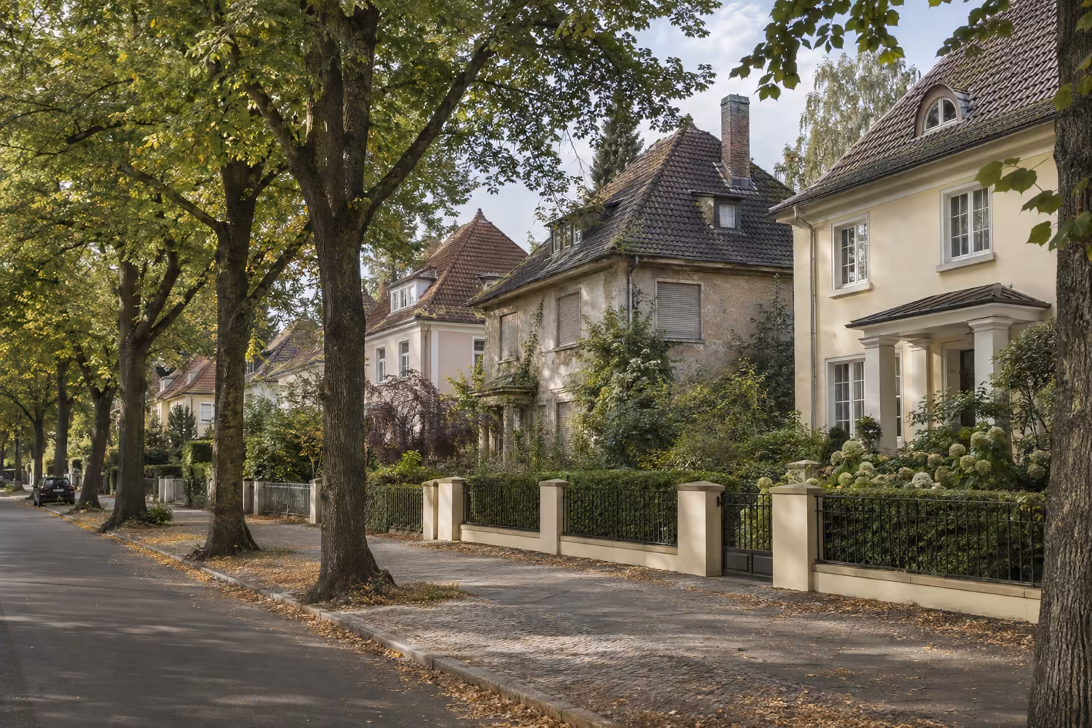 Ruhige Villenstraße mit alten Linden und Villen der 1920er und 1930er Jahre mit ähnlichen Dächern, eine davon steht sichtbar leer.