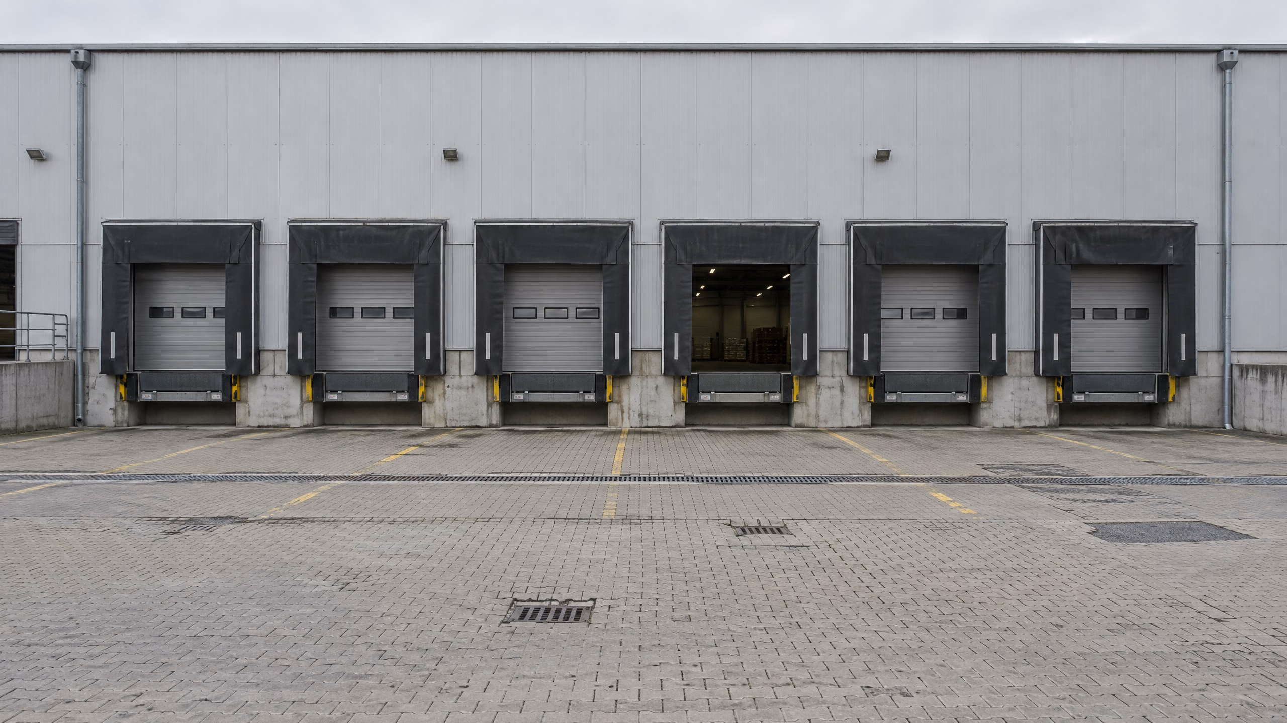 Rampenhof einer Logistikhalle: sechs Sektionaltore mit Torabdichtungen und Überladebrücken, eines offen, davor ein gepflasterter Hof mit Entwässerungsrinne, Abläufen und abgesackten Stellen.