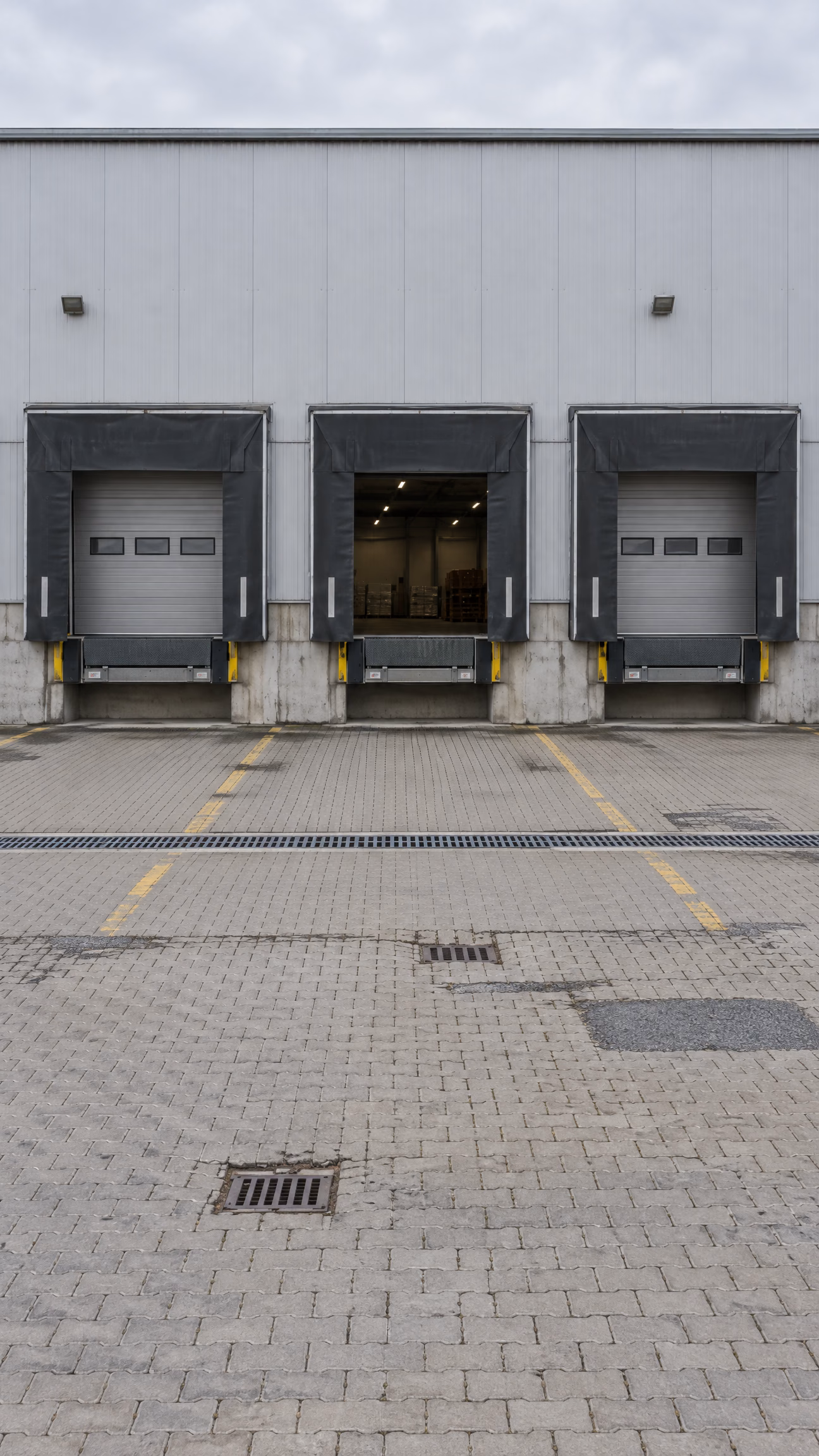 Rampenhof einer Logistikhalle: drei Sektionaltore mit Torabdichtungen und Überladebrücken, das mittlere offen, davor ein gepflasterter Hof mit Entwässerungsrinne und Abläufen.