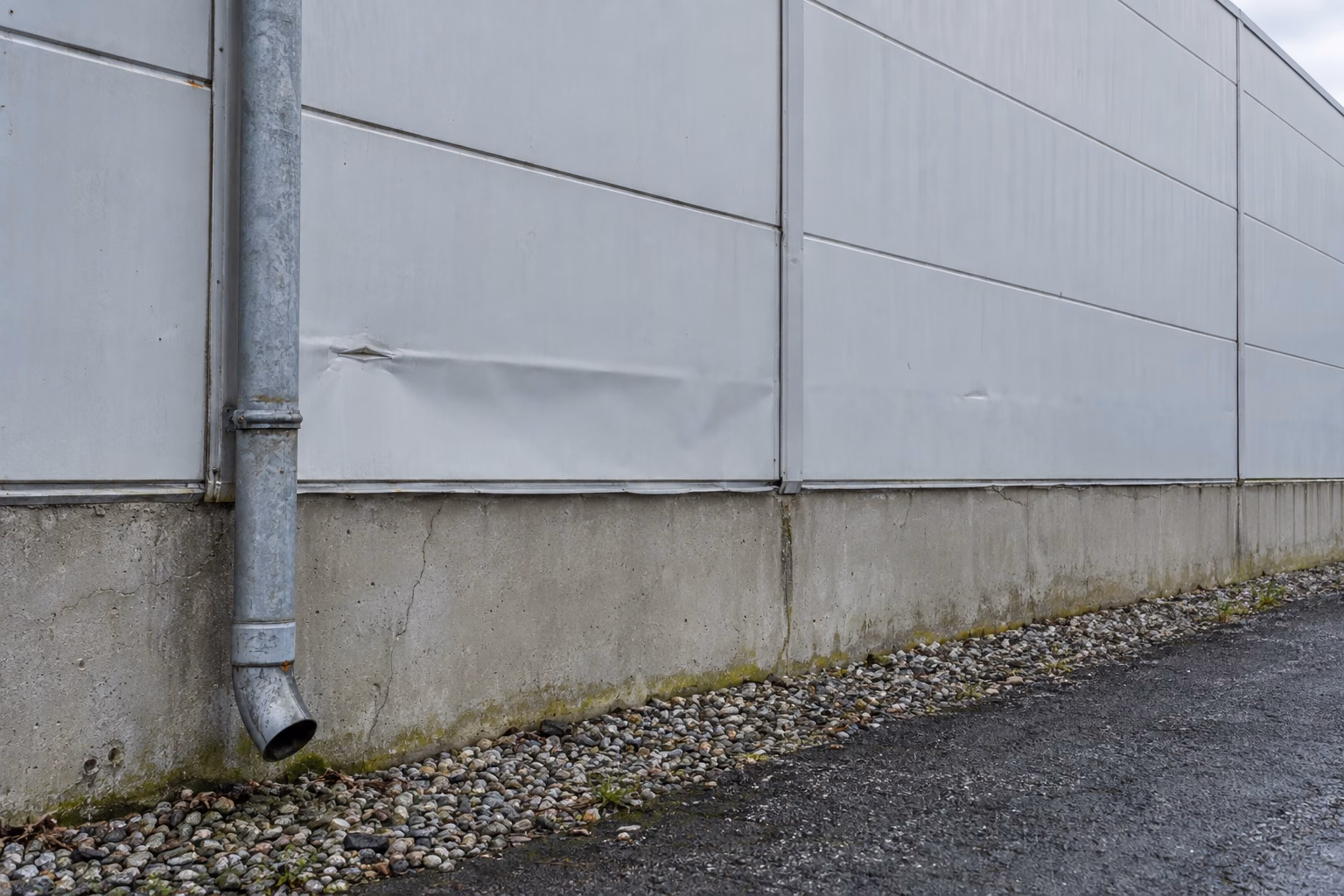 Außenwand einer älteren Lagerhalle aus Sandwichpaneelen mit verwitterten Fugen und Dellen, ein Fallrohr und ein Betonsockel mit feinen Rissen und Moos.