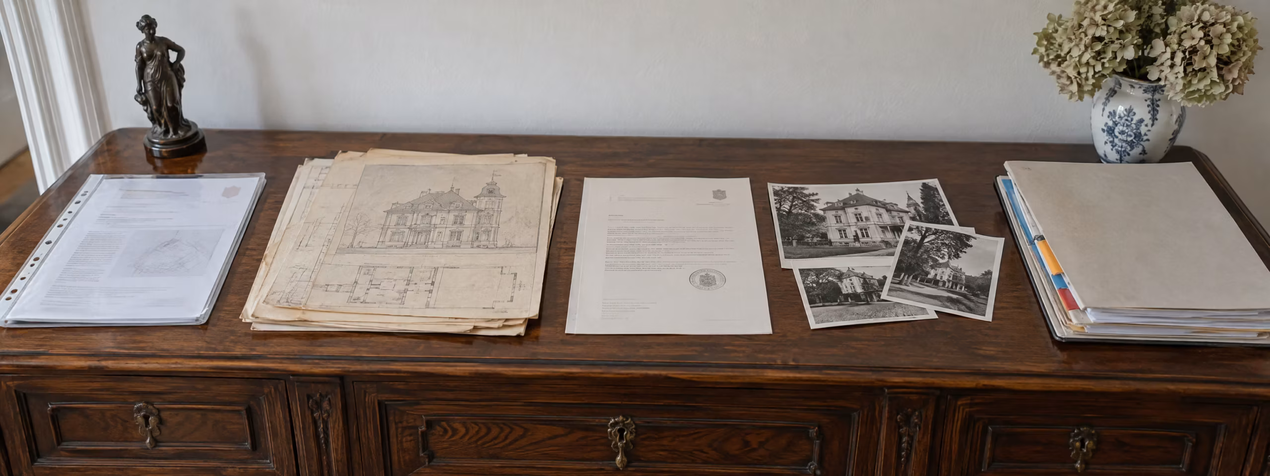Auf einem alten Holzsideboard liegen nebeneinander ein Grundbuchauszug, alte Grundrisse und eine Fassadenzeichnung, ein Schreiben der Denkmalbehörde mit Siegel, alte Fotos der Villa und ein Ordner mit Angeboten.