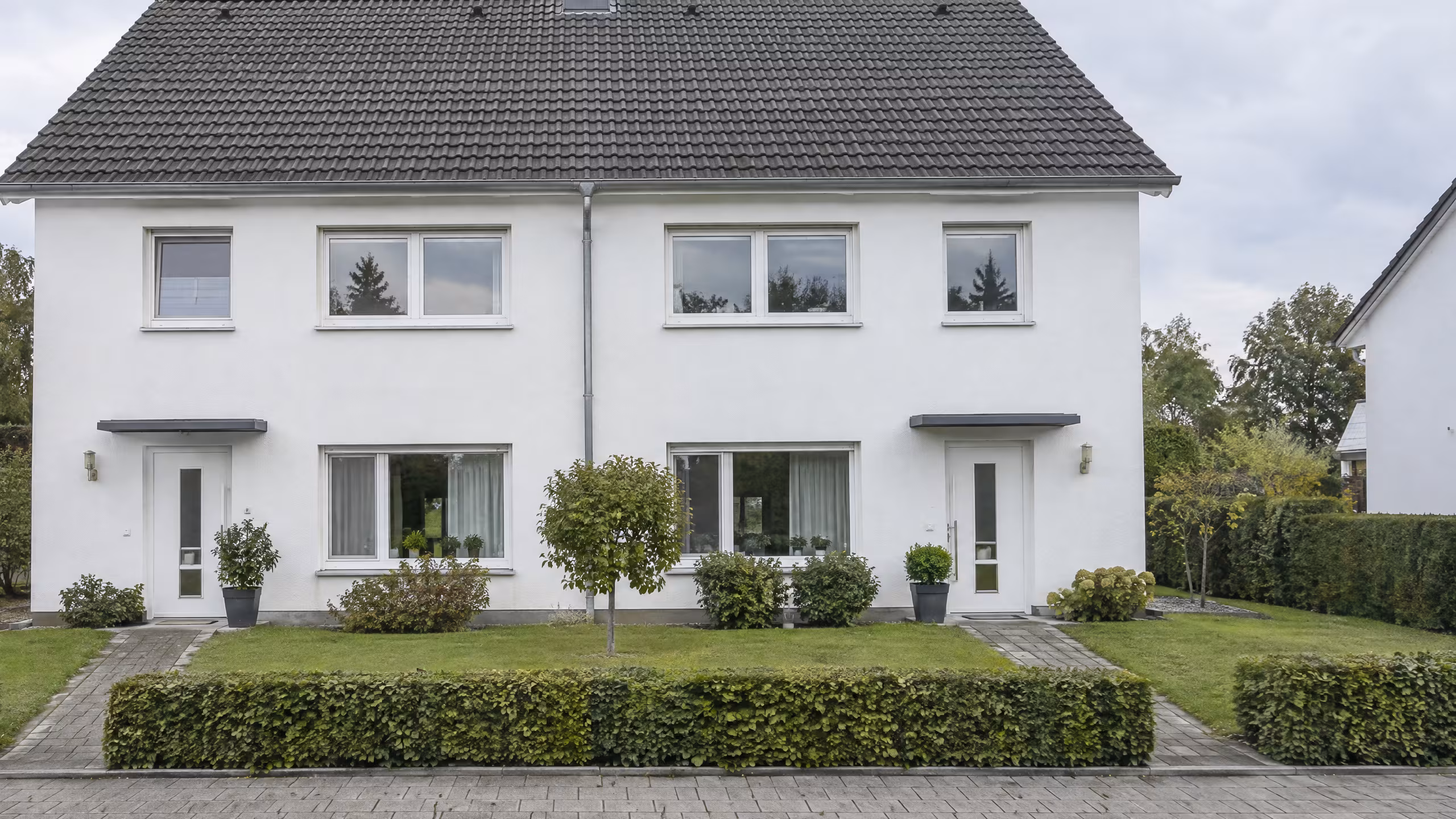Illustratives Motiv: ein gepflegtes Doppelhaus mit weißer Fassade, Rasen und Hecke unter bewölktem Himmel.