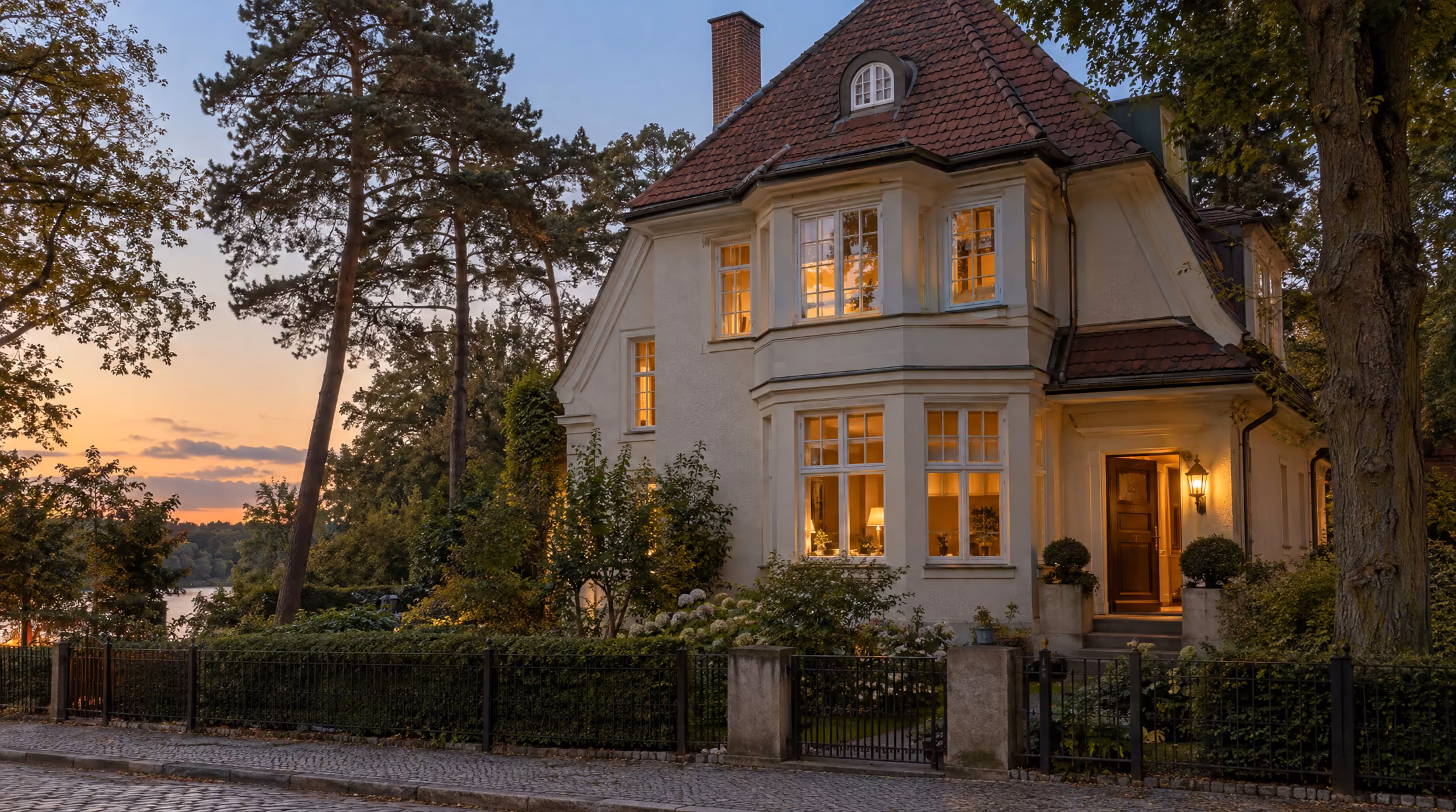 Berliner Landhausvilla am Schlachtensee im Abendlicht: cremefarbene Putzfassade mit Erker, rotbraunes Ziegeldach, beleuchtete Haustür, Eisenzaun, Kiefern und Blick auf den See.
